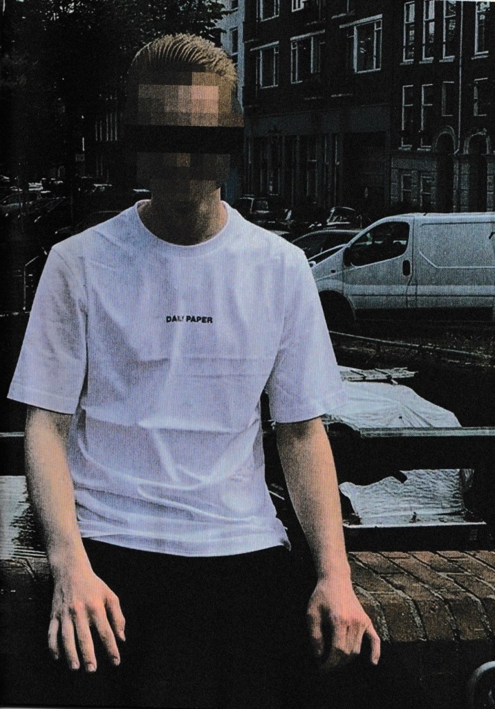 Een persoon met blonde haren, gekleed in een wit T-shirt met 'DAILY PAPER' op de borst, zit op een bankje aan een kanaal in een stadsomgeving.
